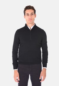 Maglione nero a manica lunga con zip a un quarto, realizzato in un tessuto liscio. Presenta un orlo e polsini a costine, indossato sopra una camicia bianca con colletto.