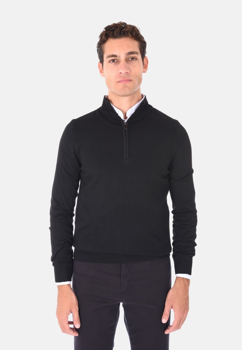 Maglione nero a manica lunga con zip a un quarto, realizzato in un tessuto liscio. Presenta un orlo e polsini a costine, indossato sopra una camicia bianca con colletto.