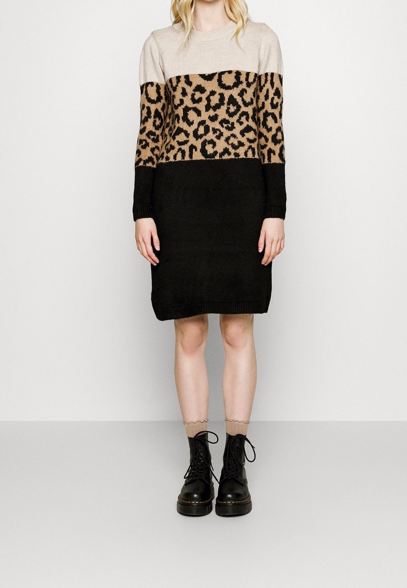 Femme portant une robe pull beige à imprimé léopard et blocs de couleur noire, avec des chaussettes marron clair et des bottes noires à lacets.