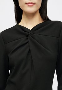 Žena nosi crni top s uvrnutim čvorom na izrezu, vidljiva su gornji dio torza i donji dio lica.