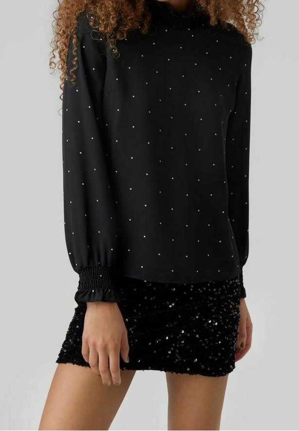 Femme portant un chemisier noir à manches longues avec des pois blancs et une mini-jupe noire à sequins, debout devant un fond uni.