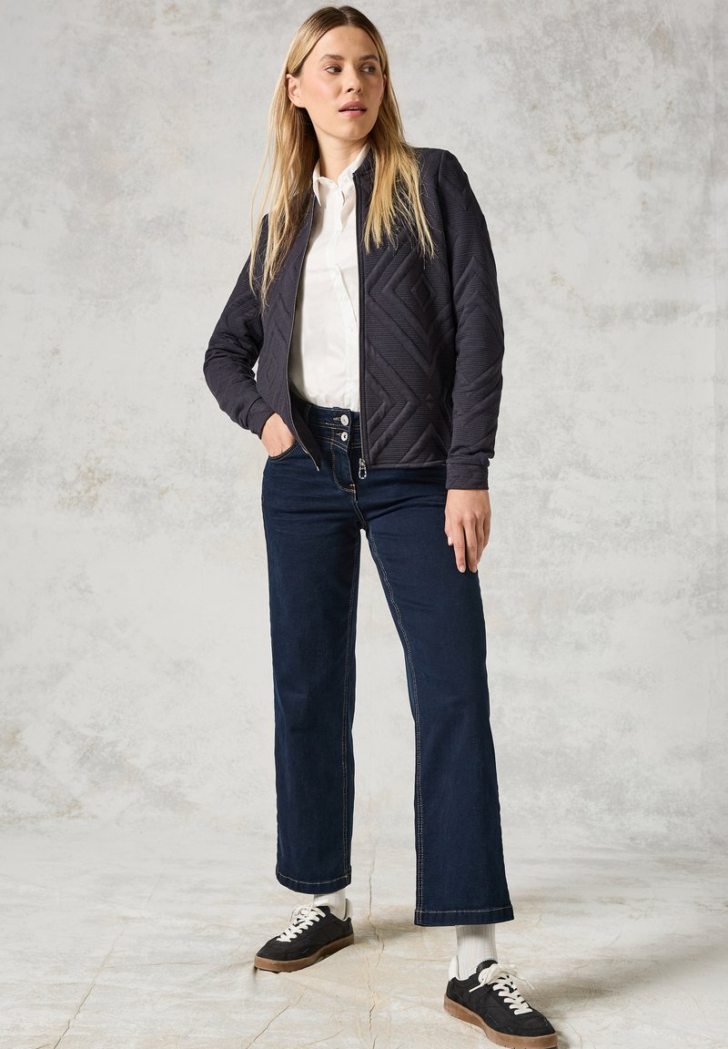 Veste matelassée bleu marine foncé avec des motifs diagonaux, fermeture éclair, accompagnée de pantalons en denim foncé évasés et de baskets noires avec des accents blancs.