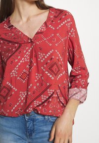 Blouse rouge à motif avec un col en V, des manches trois-quarts ornées d'accents floraux, et des motifs complexes blancs et orange partout.