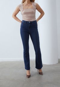 Beige formsydd topp med fyrkantig halsringning och draperade detaljer, kombinerad med mörkt blå utsvängda jeans. Modellen bär bruna spetsiga klackar. Minimalistisk miljö.