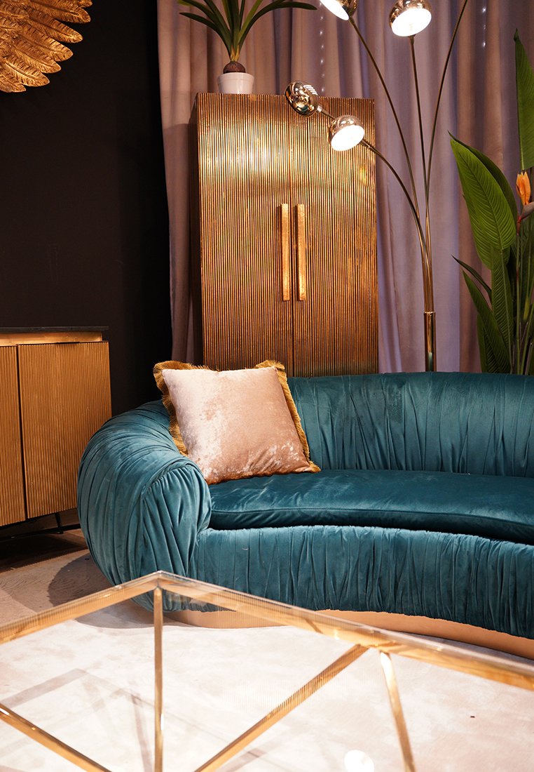 Divano in velluto teal con texture plissettata e cuscino con dettaglio dorato. Armadio a strisce dorate e lampada da terra moderna. Tavolino da caffè in vetro con base geometrica.