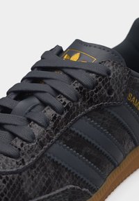 Közeli kép egy fekete Adidas Samba sportcipőről, melynek texturált kígyóbőr mintája van, fekete fűzőkkel, arany színű logóval a nyelven, és gumitalppal.