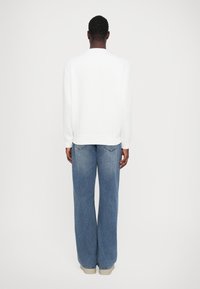 Sudadera blanca de manga larga, con cuello redondo, combinada con vaqueros de pierna ancha de mezclilla azul y zapatillas de color claro, vista desde la parte posterior.