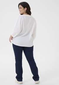 Weiße, strukturierte Bluse mit langen Ärmeln und gepufften Schultern; kombiniert mit dunkelblauen, schmal geschnittenen Jeans und hellen Schuhen.