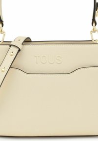 Beżowa torebka o fakturze z czarnymi wykończeniami, z frontową kieszenią i wytłoczonym logo "TOUS". Regulowany pasek z złotymi akcentami.