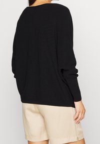 Schwarzer Strickpullover mit lockerer Passform, weitem Ausschnitt und langen Ärmeln. Kombiniert mit beigen Shorts, die einen strukturierten Stoff und eine lässige Form zeigen.