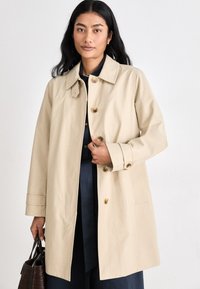 Beige trench coat with a collared neck, button closure, and two side pockets. Worn over a navy top, held with a dark handbag.