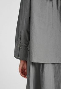 Una camicia grigia con maniche lunghe e dettagli con bottoni, dalla texture liscia e una vestibilità comoda, che copre parzialmente una gonna grigia abbinata.
