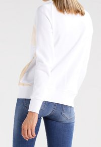 Sudadera blanca y beige con un corte relajado, puños y dobladillo acanalados, mostrada con jeans de mezclilla azul claro, centrándose en el diseño de la parte trasera.