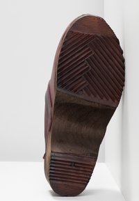 Semelle en cuir marron usée avec un talon en caoutchouc strié et une semelle à motif, reposant verticalement contre un mur et une surface blancs.