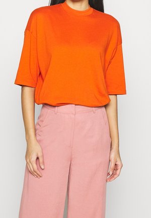 Kvinde iført en lys orange kortærmet skjorte stoppet ned i højhælede, løstsiddende lyserøde bukser mod en ensfarvet baggrund.