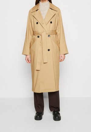 Femme portant un trench-coat beige double boutonnage avec ceinture, associé à un pantalon foncé et des chaussures noires épaisses, debout devant un fond blanc.
