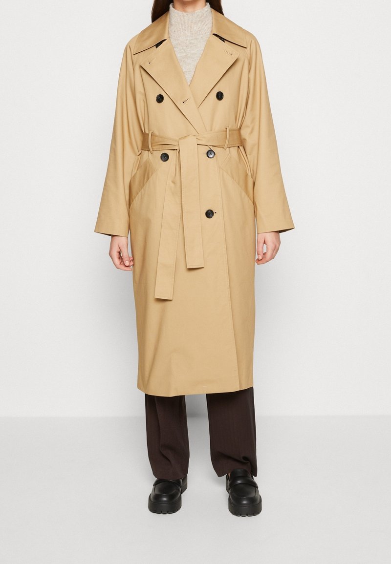 Femme portant un trench-coat beige double boutonnage avec ceinture, associé à un pantalon foncé et des chaussures noires épaisses, debout devant un fond blanc.