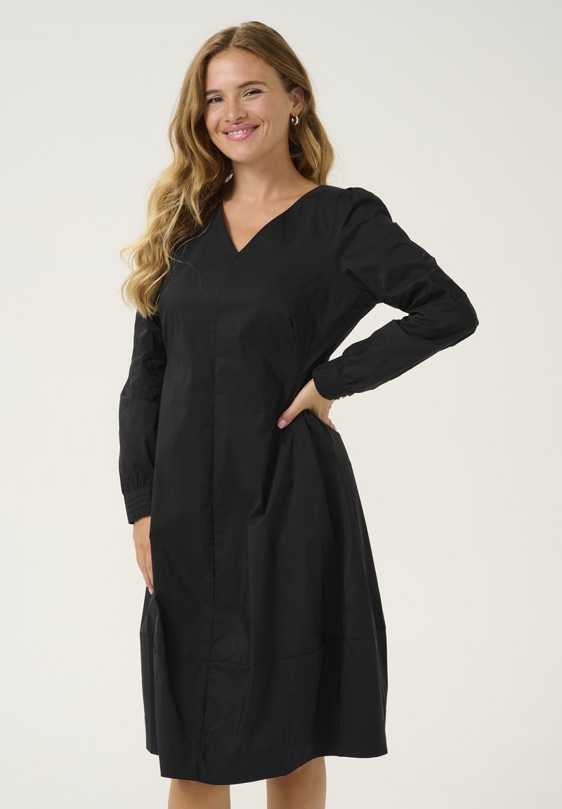 Crna haljina s V-izrezom, dugim rukavima, elastičnim manžetama i raširenim rubom. Izrađena od laganog materijala s glatkom teksturom.