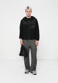 Sort fleece zip-up jakke med hætte og frontlomme, parret med grå cargo bukser og grå sneakers. Holder en lille sort taske.
