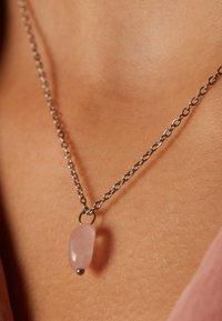 Zilveren ketting met een glad, langwerpig rozenkwarts hanger met een metalen accent aan de onderkant, rustend op de huid.