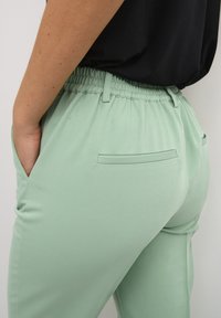 Pantaloni verde menta con vita elasticizzata, tasche laterali e una texture elegante e liscia mostrati nella vista laterale.