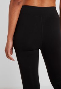 Kobieta w czarnych legginsach z wysokim stanem, widok z tyłu, z ręką luźno opadającą wzdłuż ciała na jednolitym jasnym tle.