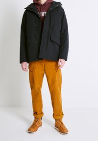 Chaqueta negra aislante con capucha, sudadera burdeos debajo, combinada con pantalones de pana naranja y botas de cuero claras con cordones.