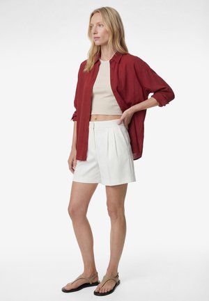 Donna che indossa una camicia rossa larga con bottoni sopra un top corto beige, pantaloncini bianchi plissettati e sandali beige, in piedi con una mano sui fianchi.