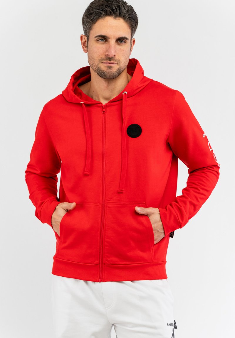 Rote Zip-Up-Hoodie mit einer Kapuze mit Kordelzug, Fronttaschen und einem schwarzen runden Logo-Patch auf der Brust. Glatter Stoff, taillierte Passform.