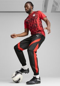 Rød og svart fotballdress med geometriske mønstre på skjorten, kombinert med svarte bukser med røde striper og svarte fotballsko.