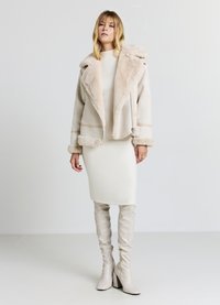 Giacca in pelliccia sintetica beige con ampio colletto, chiusura con zip e polsini coordinati. Indossata sopra un vestito bianco attillato e stivali bianchi al ginocchio.