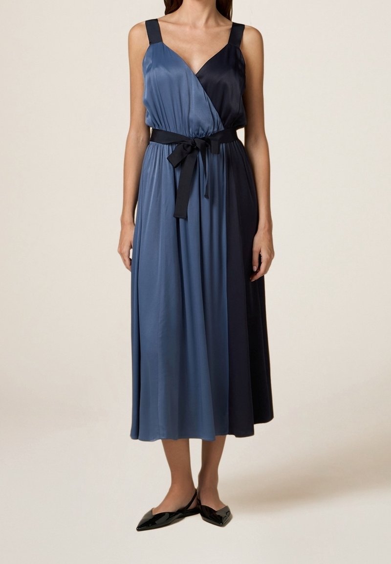 Ärmelloses Wickelkleid mit blauen und dunkelblauen Paneelen, einem Bindegürtel in der Taille und glatter, satinähnlicher Textur. Ballerinas ergänzen den Look.