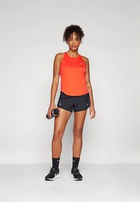 Punane sporditopp, mustad lühikesed püksid, hoiab käes veepudelit, määratud mustade tossude ja sokkidega. Lühikesed, lokkis juuksed; seisab neutraalsel taustal.