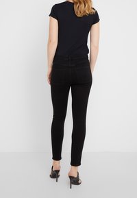 Nainen seisoo selin mustissa slim-fit-farkuissa, mustassa lyhythihaisessa paidassa ja mustissa korkokengissä valkoista taustaa vasten.