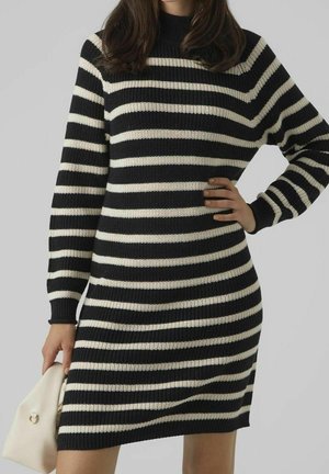Femme portant une robe en maille à manches longues rayée noir et blanc horizontale, tenant un petit sac à main beige sur un fond uni.