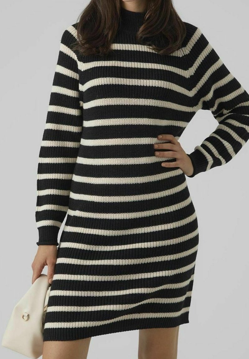Femme portant une robe en maille à manches longues rayée noir et blanc horizontale, tenant un petit sac à main beige sur un fond uni.