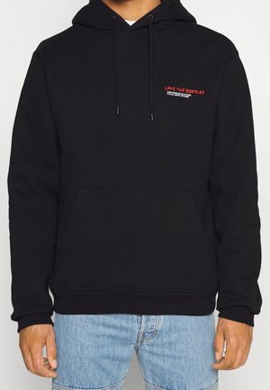 Černá mikina s malým červeným a bílým nápisem "Love The Kooples" na hrudi, nošená osobou v modrých džínách, ruce volně podél těla.