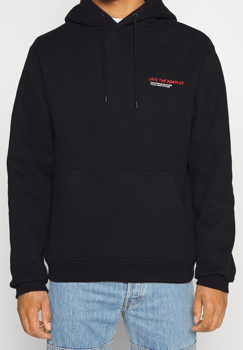 Sweat à capuche noir avec petit texte rouge et blanc "Love The Kooples" sur la poitrine, porté par une personne en jean bleu, mains détendues le long du corps.