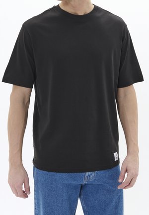 Mand iført en ensfarvet sort kortærmet T-shirt og blå jeans, stående med armene afslappet ned langs siderne mod en hvid baggrund.