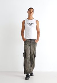 Un débardeur blanc sans manches avec un design graphique noir, associé à un pantalon cargo vert olive ample avec plusieurs poches et une coupe décontractée.
