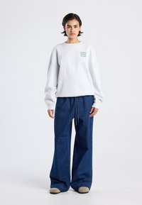 Joven mujer de pie con la mirada al frente, vestida con una sudadera blanca con el texto "Sunday Coffee Club" y unos vaqueros de pierna ancha azules, acompañados de zapatos beige.