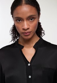 Blusa nera in seta con colletto, bottoni frontali e una singola tasca sul petto. Il tessuto è liscio con una leggera lucentezza.