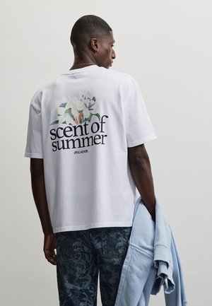 Man in wit T-shirt met bloemmotief en tekst "geur van de zomer" op de rug, houdt een lichtblauw jasje vast, tegen een effen achtergrond.