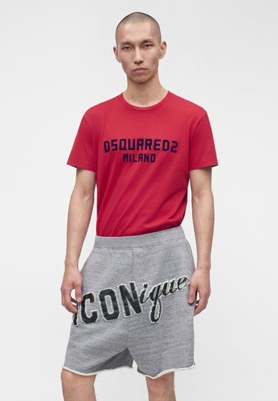 Άνδρας φοράει κόκκινο T-shirt DSQUARED2 Milano και γκρι σορτς με μεγάλο κείμενο "ICONique", στέκεται μπροστά σε απλό ανοιχτό φόντο.