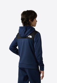 Marineblå hettegenser med hette, med et svart bærestykke og hvit logo på øvre del av ryggen. Mykt stoff med en jevn tekstur.