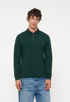 Homme aux cheveux bouclés portant un polo manches longues vert foncé avec fermeture éclair et un jean bleu clair sur un fond uni clair.