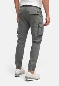 Homme portant un pantalon de jogging cargo gris avec poches à rabat et des baskets blanches New Balance, debout devant un fond uni.