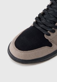 Κοντινό πλάνο στη μύτη ενός μαύρου και μπεζ αθλητικού παπουτσιού με διάτρητο μαύρο σουέντ και μαύρα κορδόνια με λευκή ραφή.