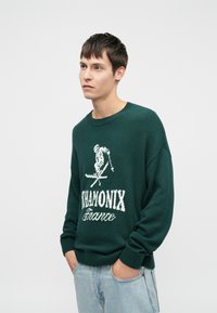 Zeleni pleteni pulover s okroglim ovratnikom in rebrastimi manšetami. Na sprednji strani ima bele grafike smučanja in besedilo "CHAMONIX Francija".