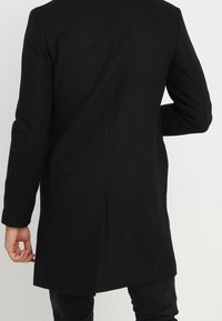 Cappotto oversize in lana nera con taglio dritto, maniche lunghe e senza bottoni o accessori visibili. Design semplice con lunghezza a metà coscia.